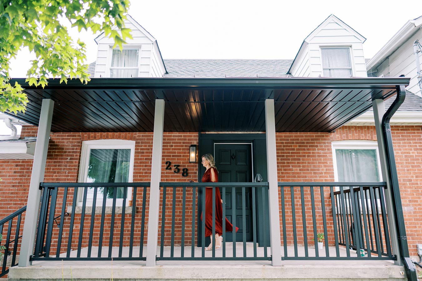 Modern brick house with black door and woman in red dress walking on covered porch, showcasing residential architecture and exterior design features.