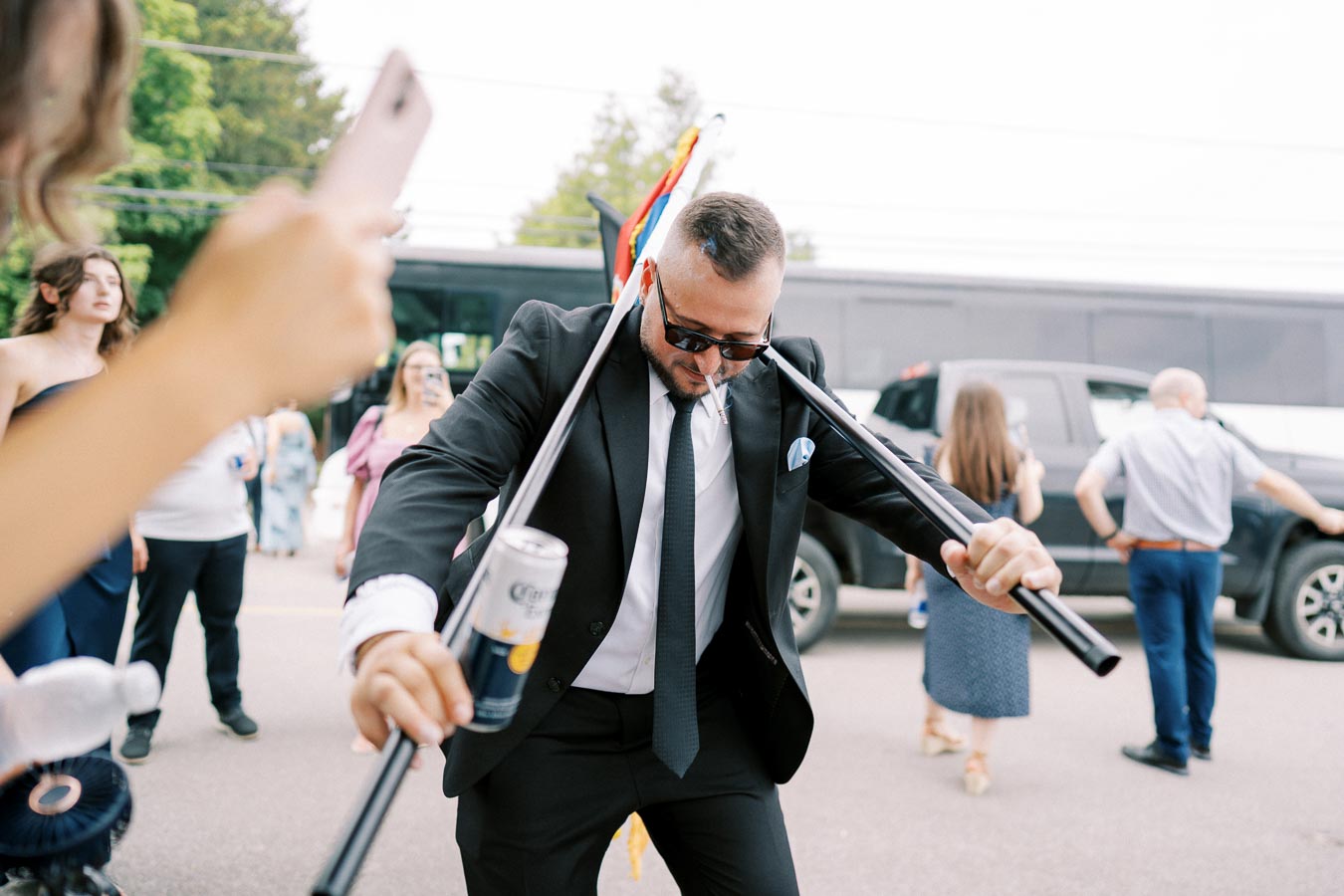 Man in a suit and sunglasses having fun at an outdoor celebration, holding a beverage and dancing energetically while people around take photos and enjoy the event.