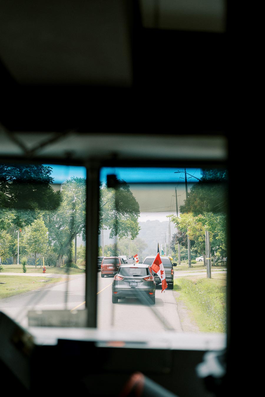 Parade of cars displaying Canadian flags on a rural road, viewed through a vehicle window, with trees lining the street under an overcast sky.