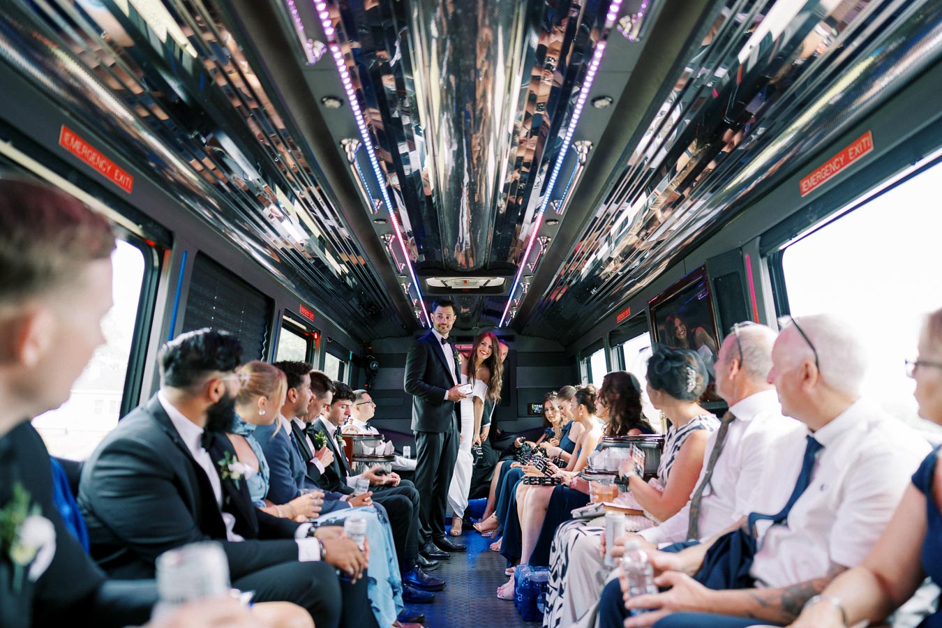 Wedding party inside a luxury limousine bus with LED lighting, celebrating with drinks, dressed in formal attire.