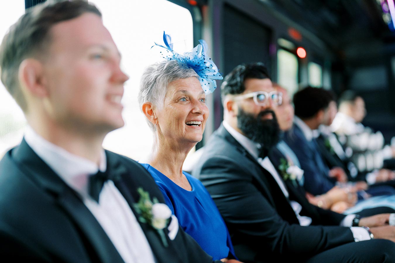 A group of elegantly dressed people sitting inside a bus, with a woman in a blue dress and fascinator smiling and sitting between two men in suits with boutonnieres, suggesting a wedding or formal event setting.