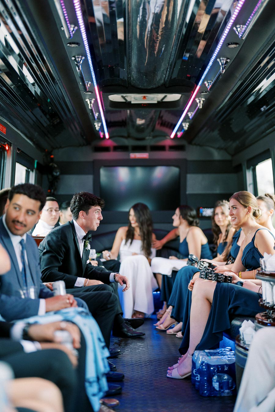 Group of elegantly dressed people inside a party bus with vibrant ceiling lights, enjoying a festive atmosphere.