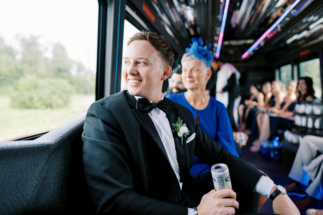 Man in a tuxedo smiling on a bus, holding a beverage can, with a woman in a blue dress and hat seated nearby, surrounded by other people.