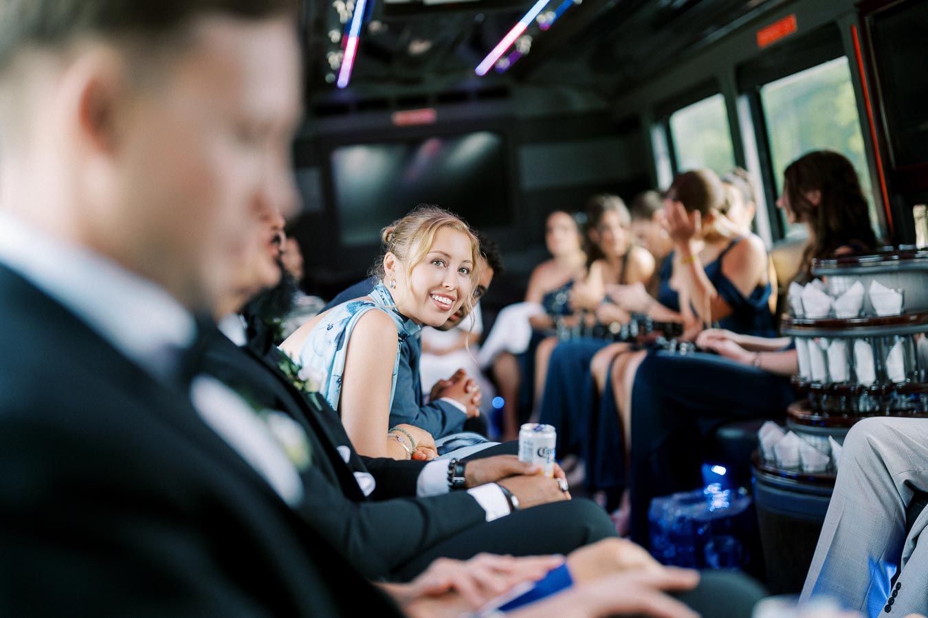 A group of people dressed formally inside a party bus, with one person smiling and holding a drink, surrounded by colorful lights.
