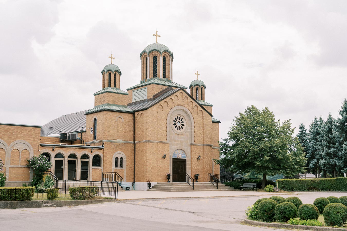 Architectural view of a traditional Orthodox church with brick walls and green-domed towers, set against a cloudy sky. The church features a large circular window above the main entrance and is surrounded by well-maintained landscaping including trees and shrubs.