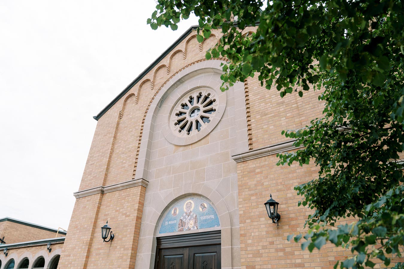 Exterior view of a historic brick church with a detailed circular stained glass window, framed by leafy green trees, showcasing classic architecture and ornate design.