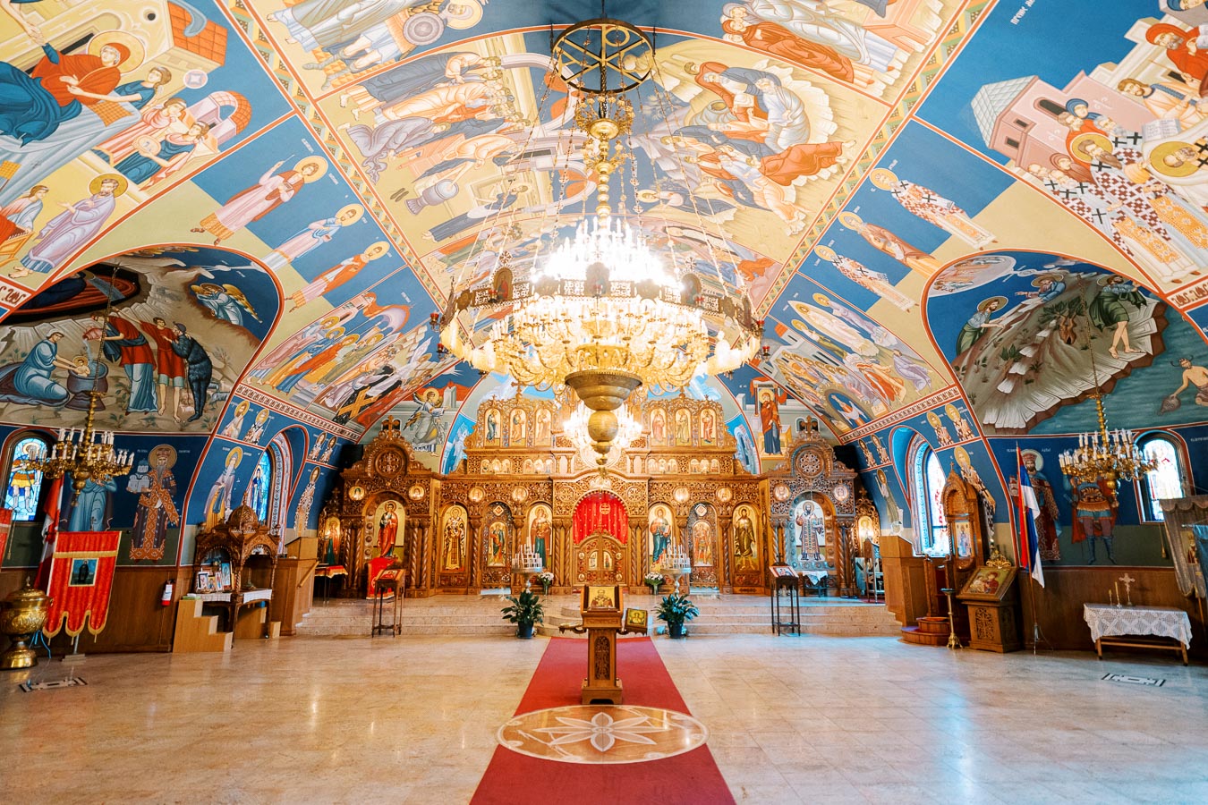 Intricately decorated interior of a Serbian Orthodox church with vibrant frescoes, ornate wooden icons, and a central chandelier, showcasing traditional religious art and architecture.