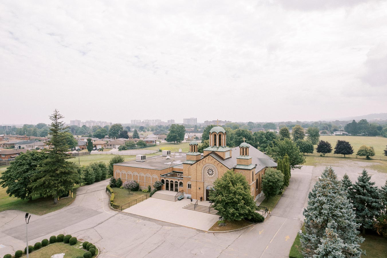 Aerial view of a historic brick church with arched windows and domed rooftops surrounded by lush greenery and trees, illustrating serene suburban scenery with houses and open fields in the background.