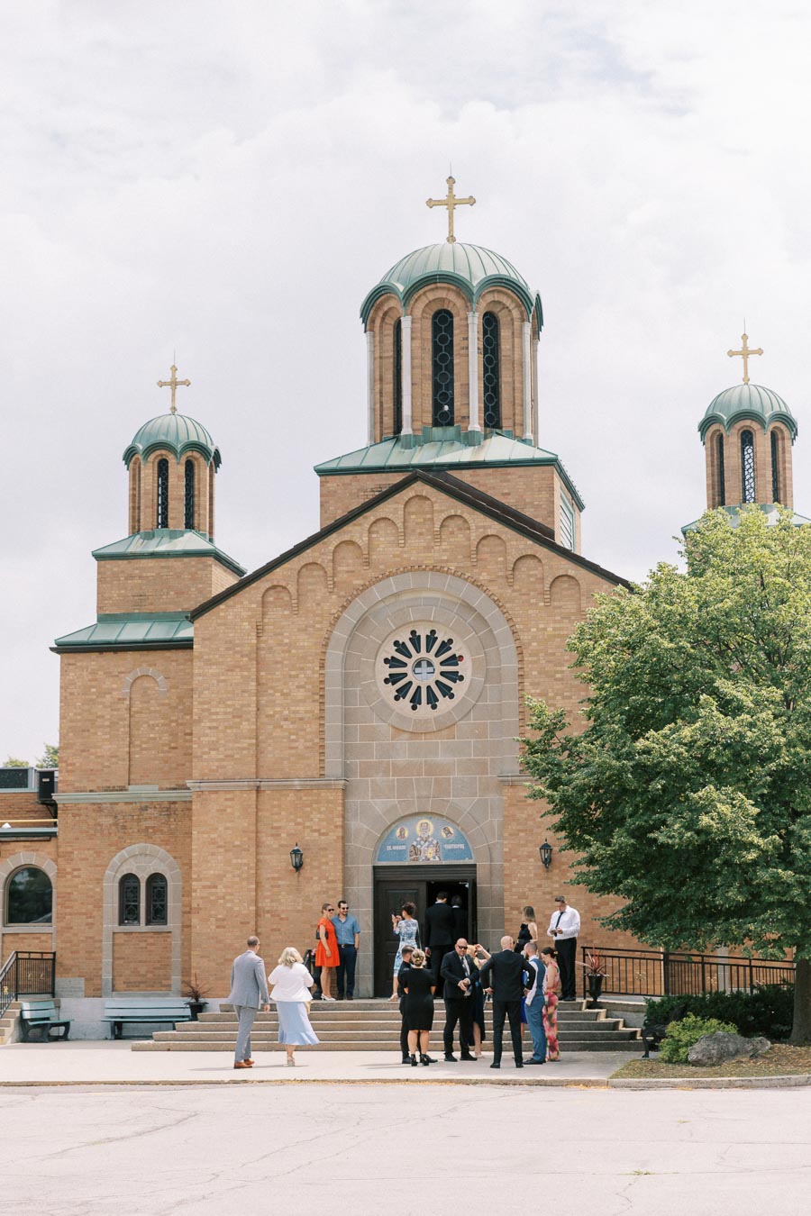 Front view of a historic brick Orthodox church with three green domes and crosses, people gathered at the entrance on a sunny day, highlighting cultural architecture and community gathering.