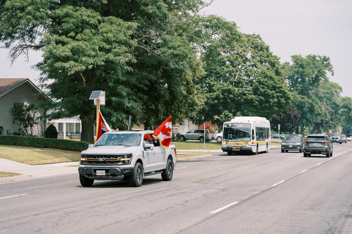 A white pickup truck displaying Canadian flags drives down a suburban street lined with trees and residential houses, followed by a public transit bus and other vehicles.
