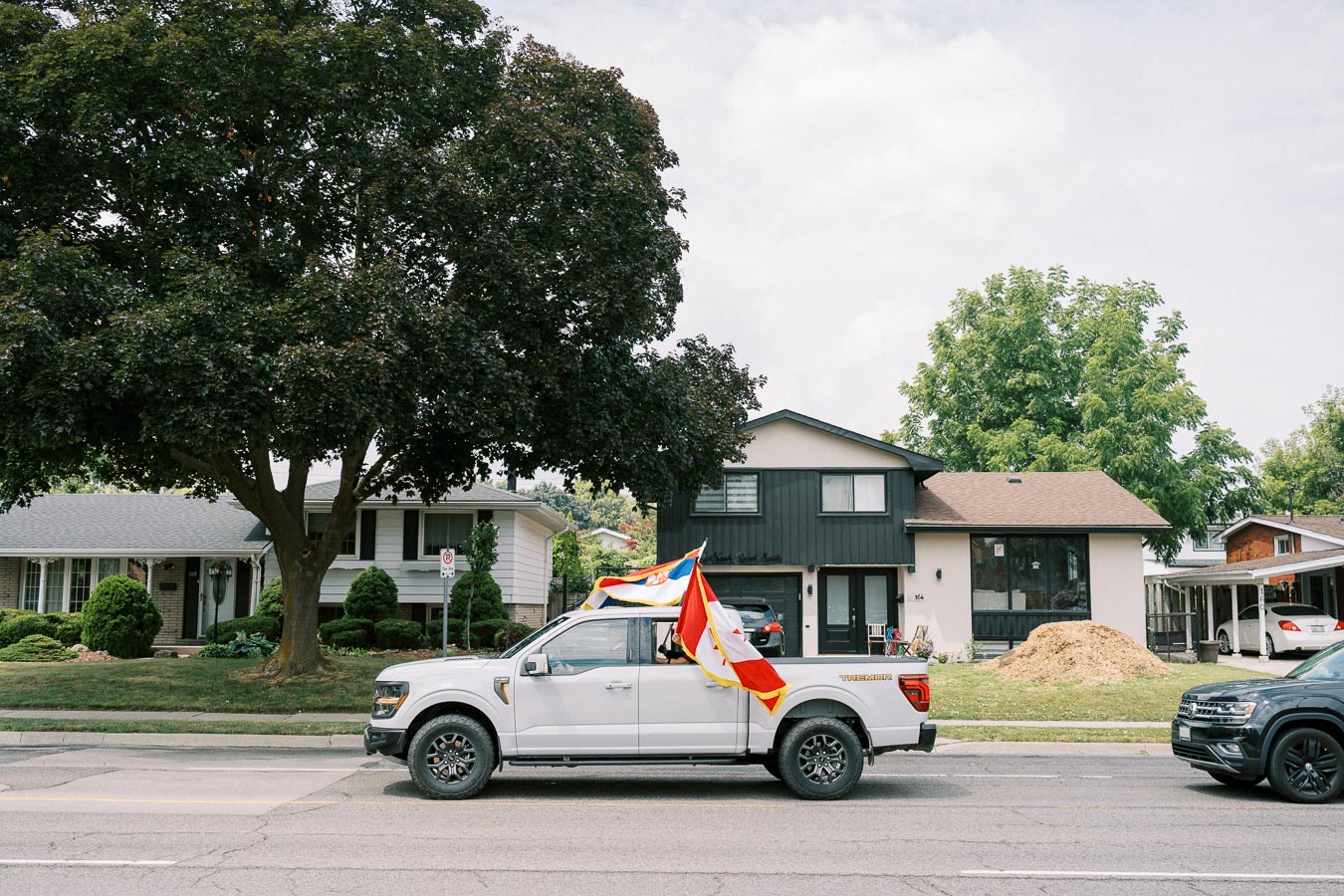 White pickup truck decorated with colorful flags driving down a residential street, with houses and trees in the background.
