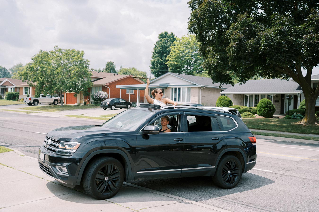 A black SUV parked on a suburban street with a smiling bride joyfully waving from the sunroof, showcasing a lively wedding celebration.