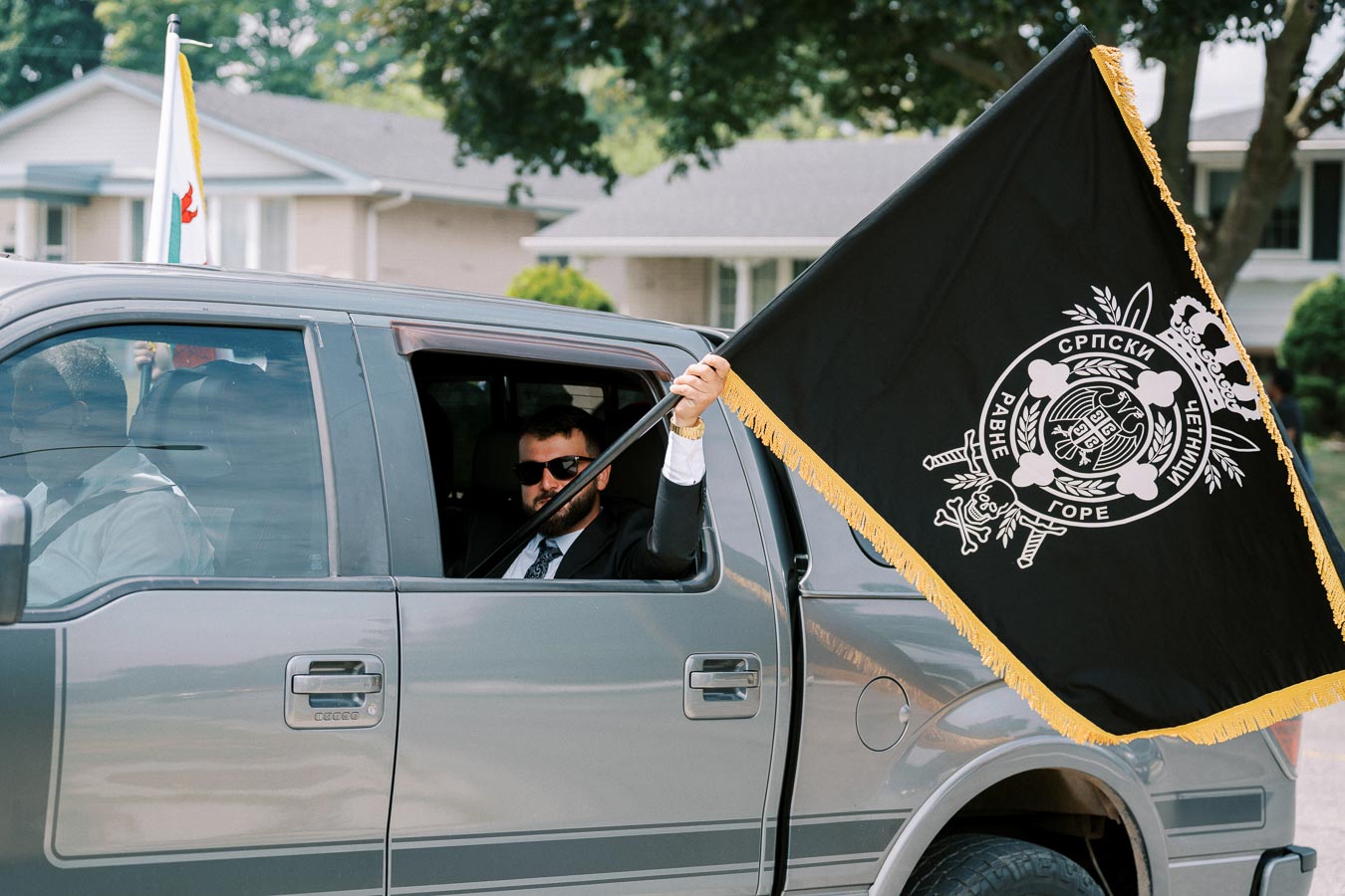A man in a suit holding a black flag with intricate emblems and text, sitting in a grey pickup truck. The vehicle is parked on a suburban street with trees and houses in the background.