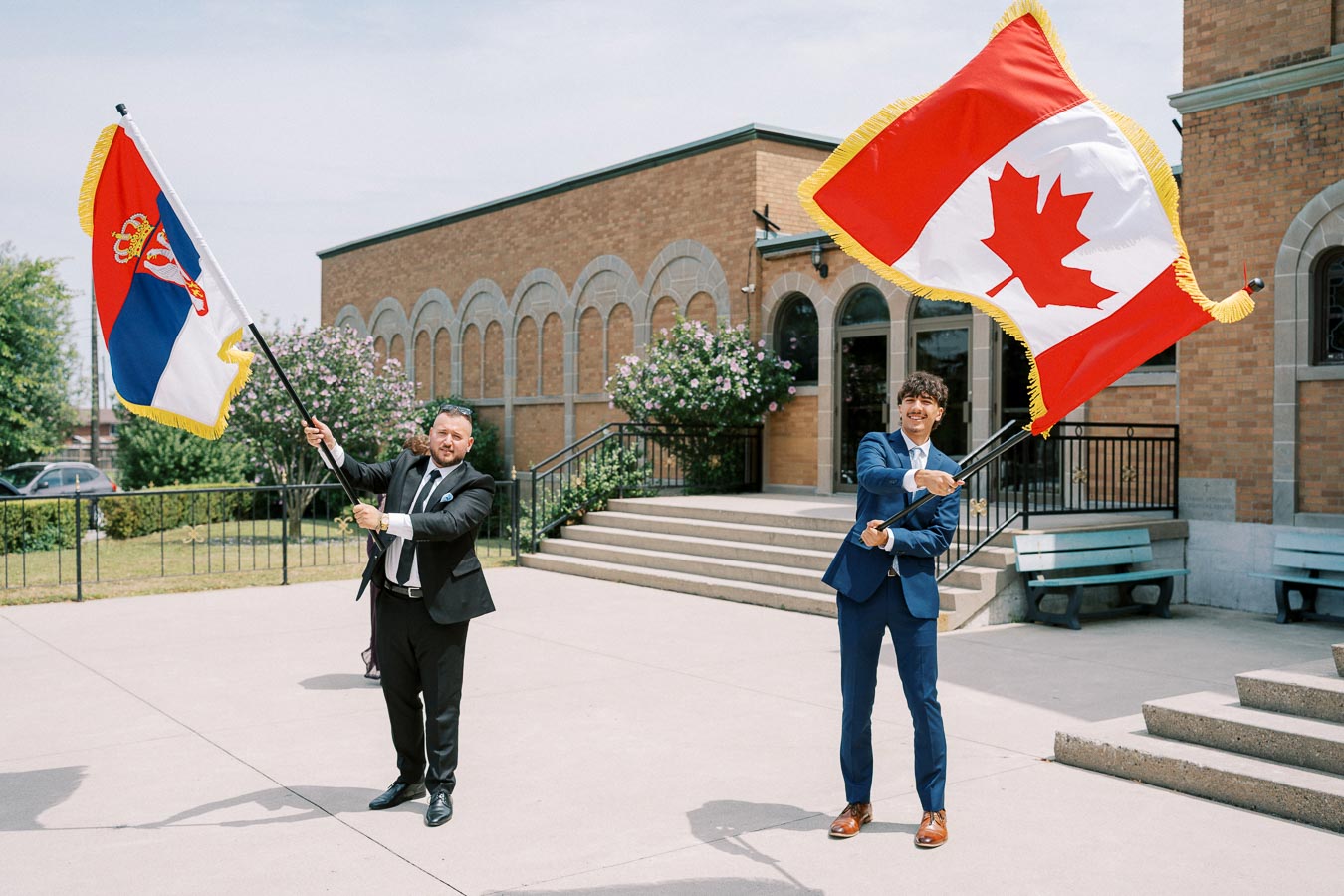 Two men in suits joyfully waving Serbian and Canadian flags outside a brick building on a sunny day.