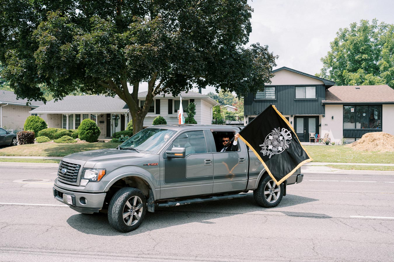 Silver Ford F-150 truck driving down a residential street with a person holding a large black flag out the window, suburban houses and green trees in the background.