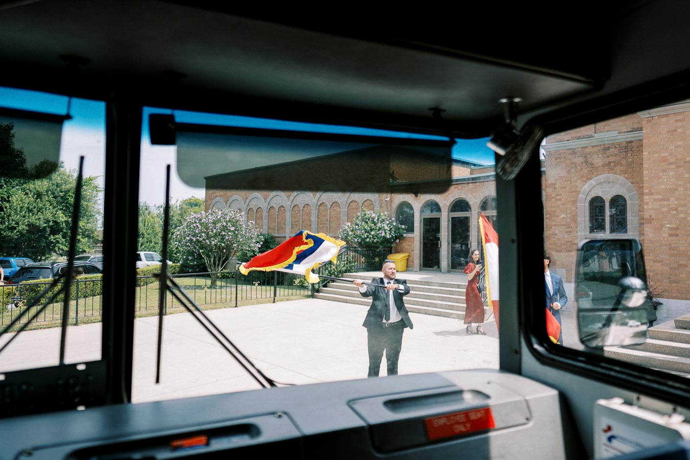 View from inside a vehicle showing a man waving a colorful flag in front of a brick building with arched windows and greenery, creating a vibrant outdoor scene.