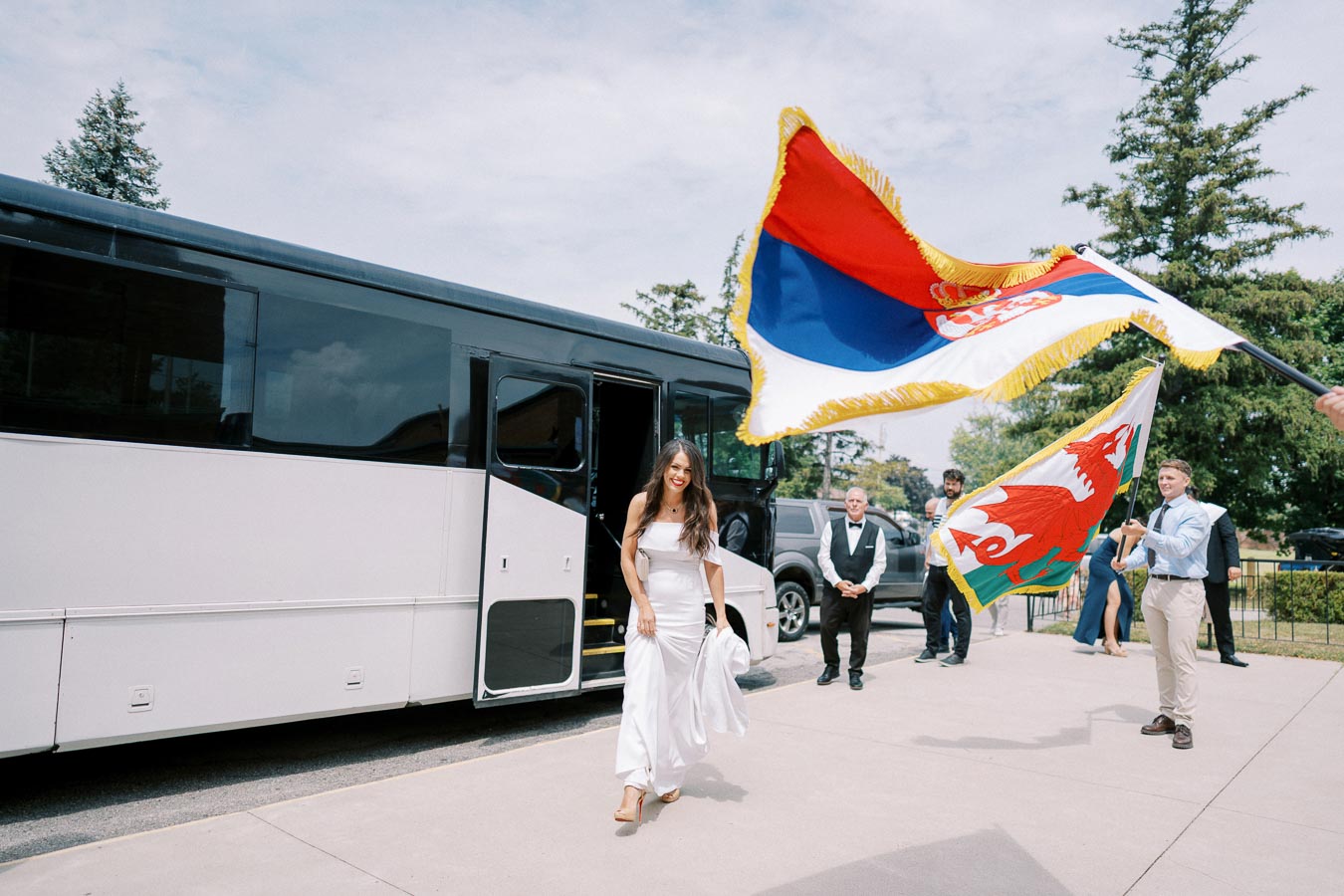 A woman in a white dress smiling while exiting a tour bus, with people holding and waving colorful flags in a festive outdoor setting, surrounded by trees and a clear sky.