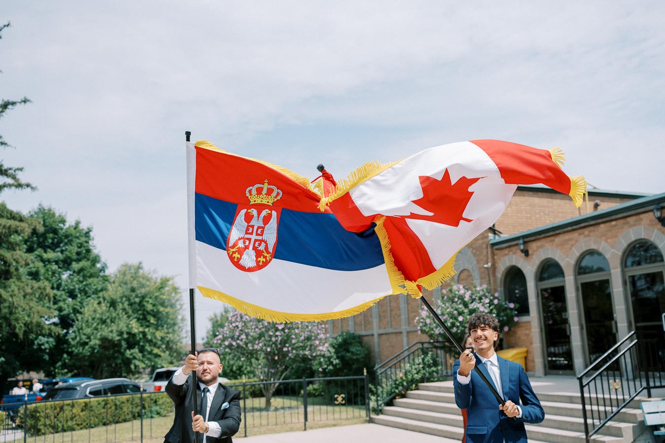 Two people holding large flags of Serbia and Canada, standing outdoors in front of a building with arched windows, under a partly cloudy sky.