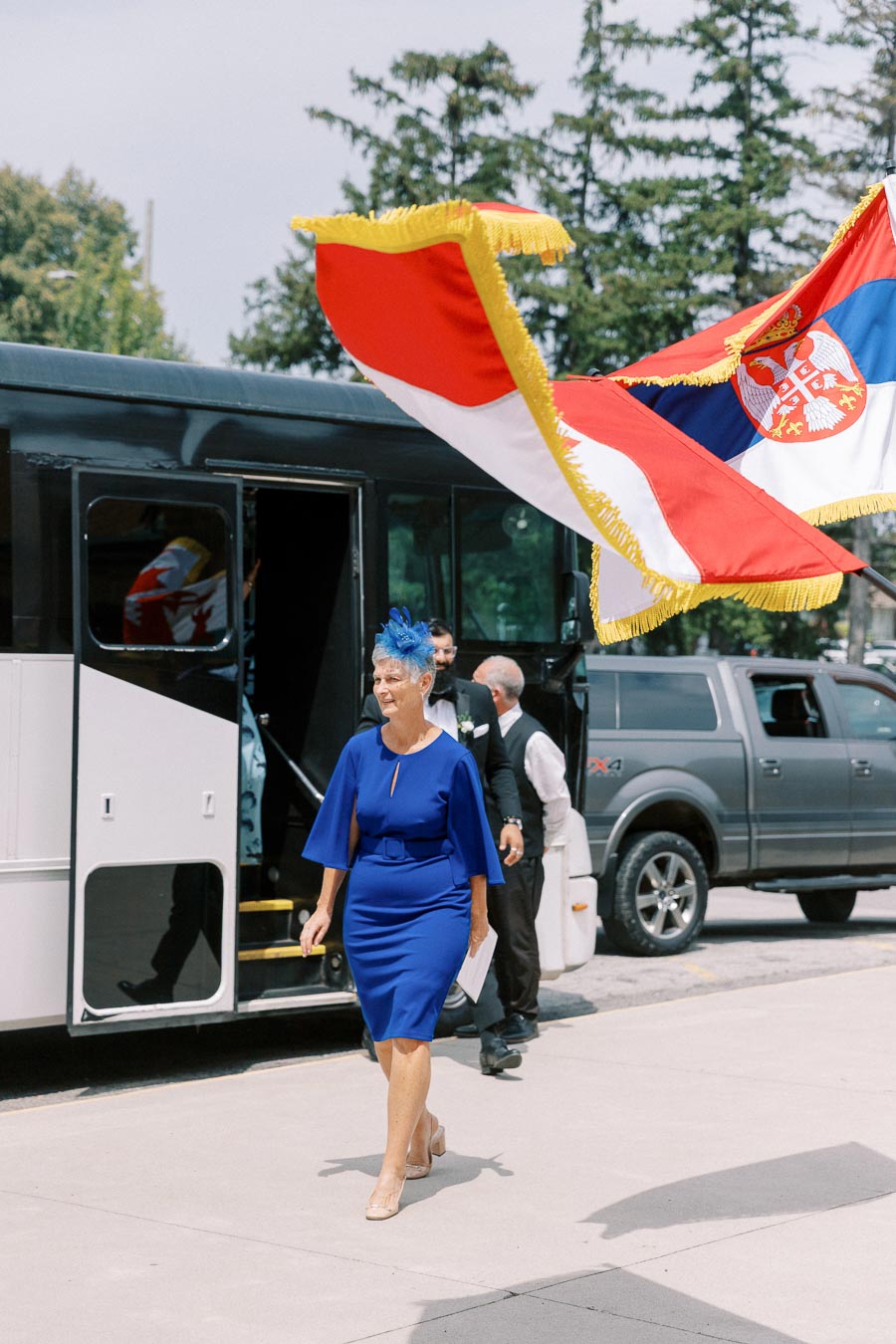 Elegant woman in a blue dress walking past a bus with a Serbian flag waving above, near parked vehicles and trees.
