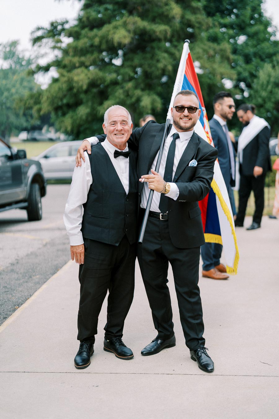 Two men in formal attire smiling outdoors, one holding a flag, with people in formal wear in the background.