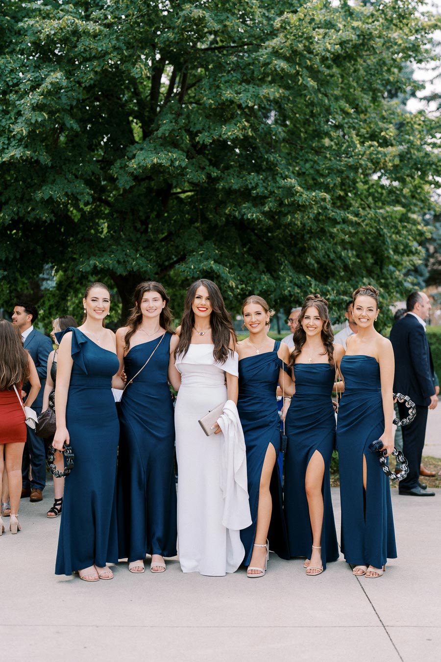 A group of six women posing outdoors, five wearing matching navy blue dresses with high slits, and one in a white dress, standing on a paved walkway with a lush green tree in the background.