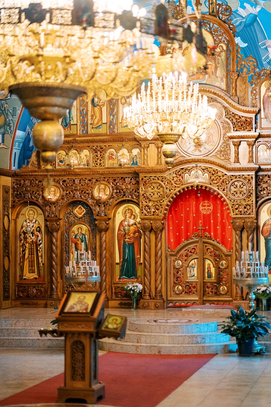Ornate interior of an Eastern Orthodox church featuring intricate wooden iconostasis, vibrant religious icons, chandeliers, and a red carpet leading to the altar.
