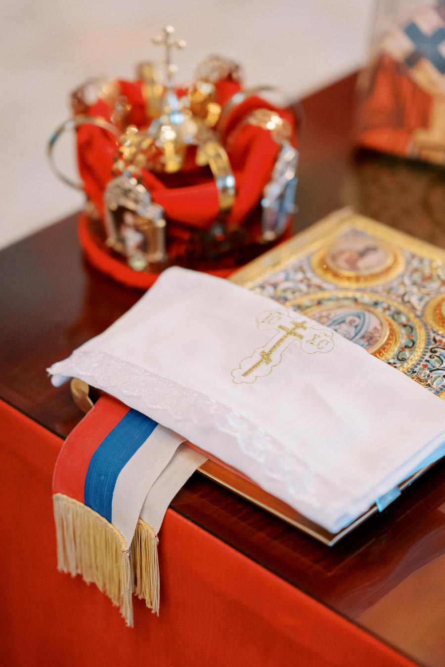 Ceremonial religious items on a table, including an ornate crown, a cloth with a golden cross, and a richly decorated book. The cloth includes red, blue, and white ribbons. Perfect for an Orthodox wedding or religious event.