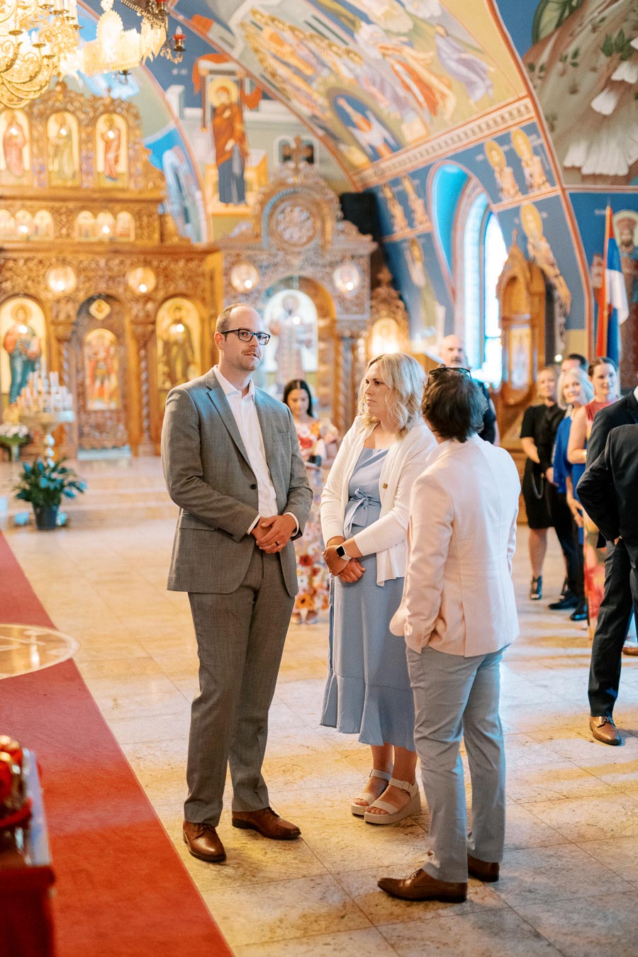 A group of formally dressed people standing inside an ornately decorated church with colorful murals and icons, engaging in conversation during a social gathering or event.