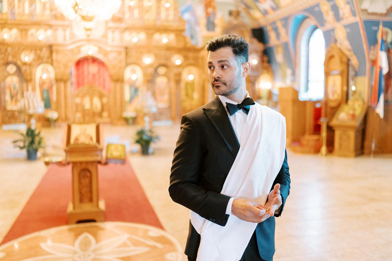 A man in formal attire with a black suit and bow tie, holding a white draped cloth, stands in the ornate interior of a richly decorated church, featuring golden icons, chandeliers, and a red carpet.