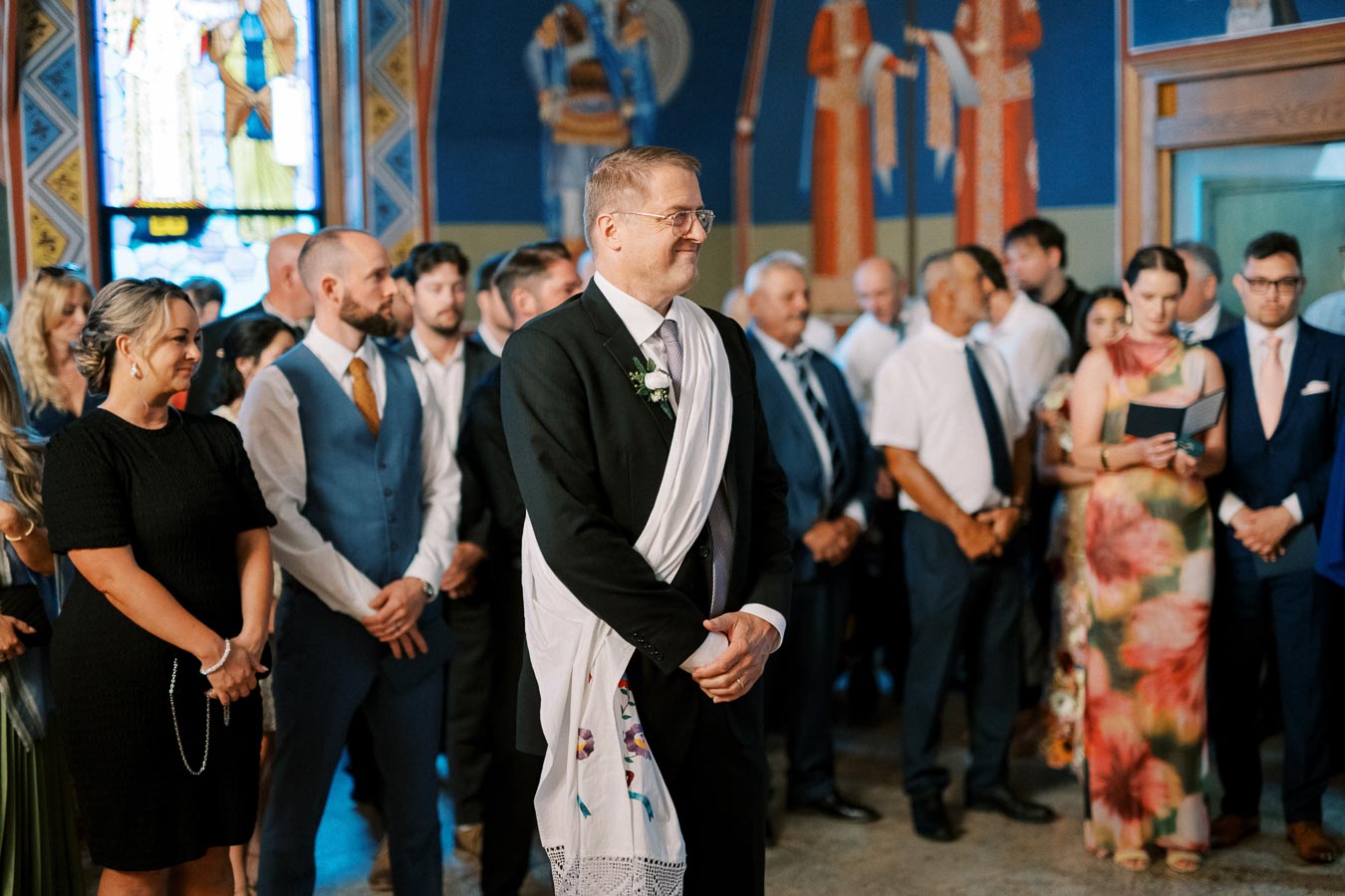 A groom stands smiling at the altar during a wedding ceremony, surrounded by elegantly dressed guests in a beautifully decorated church with vibrant stained glass windows and murals.