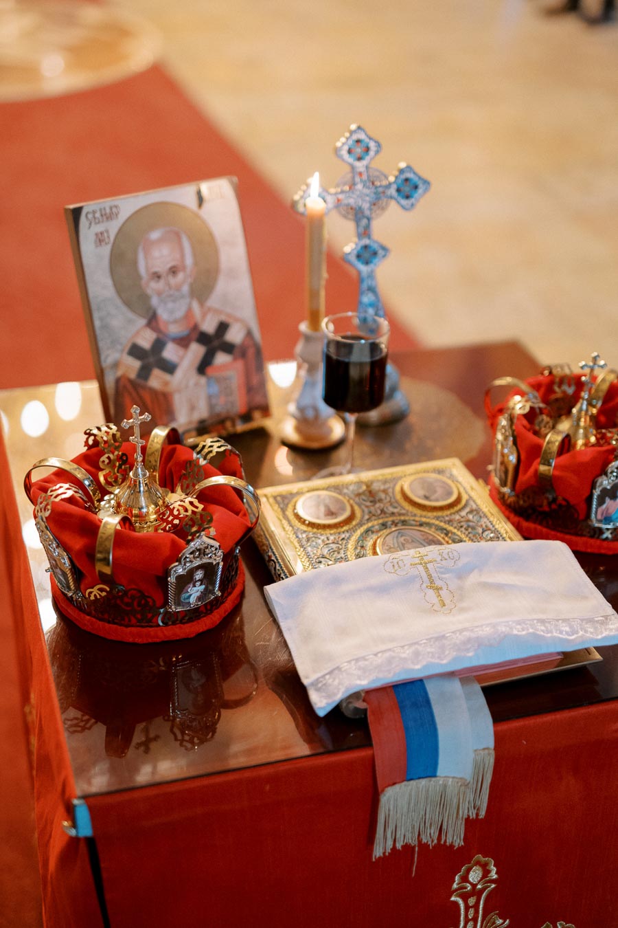 A church altar setup with religious items for a ceremony, featuring a traditional icon, two ornate crowns with red fabric, a decorative cross, a candle, a glass of wine, and an embellished book. The scene is set on a red cloth, highlighting the spiritual significance and solemnity of the occasion.