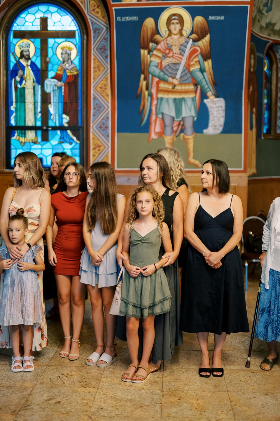 A group of women and girls standing together in a church with colorful stained glass windows and religious murals in the background.
