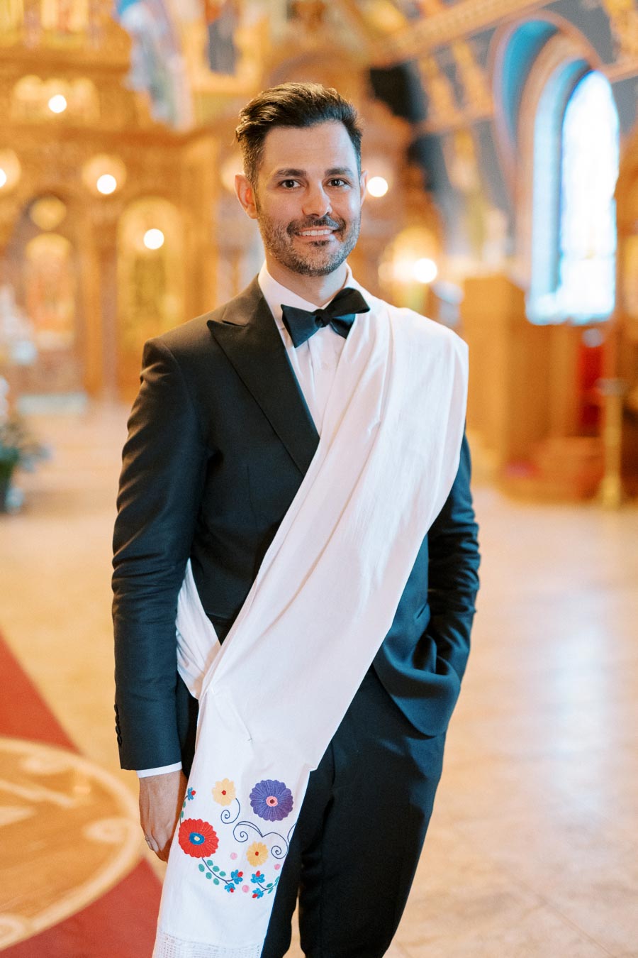 A man in a formal black suit and bow tie, draped with a white sash adorned with colorful floral embroidery, stands confidently in an ornately decorated, warmly lit interior, possibly a ceremonial venue or church.