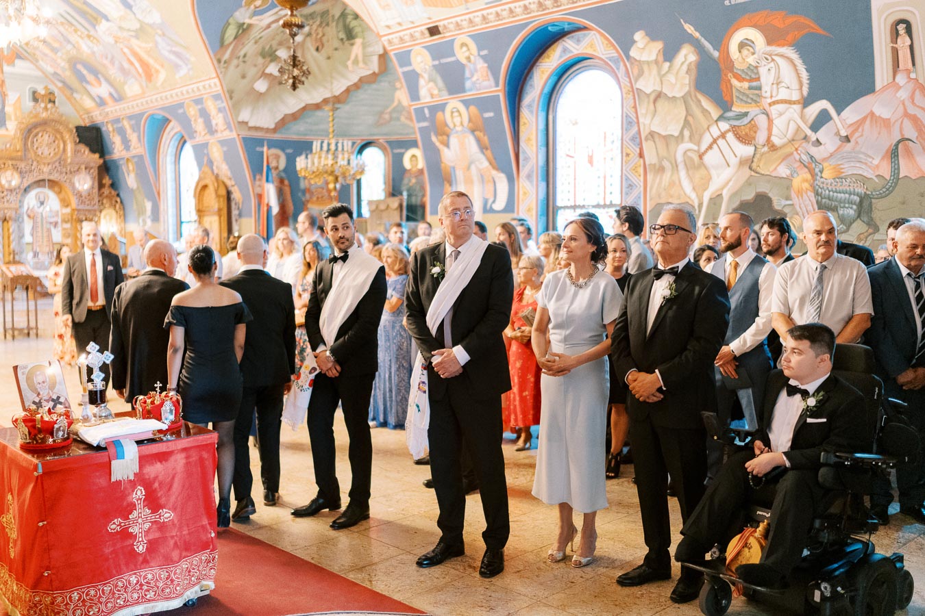 A large group of people attending a formal event inside a beautifully decorated church, with ornate murals and arched windows. The attendees are dressed in elegant attire, including suits and dresses. In the foreground, a table draped with a red cloth displays ceremonial items, including crowns and a decorative cross.