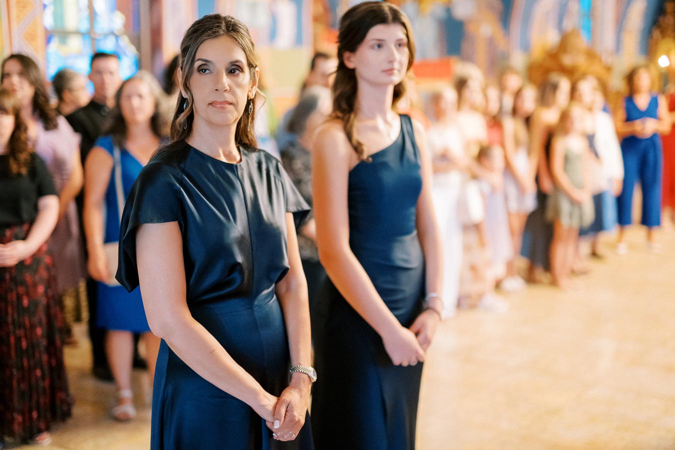 A group of elegantly dressed people gathered in a brightly lit venue, with two women in navy blue dresses standing prominently in the foreground.