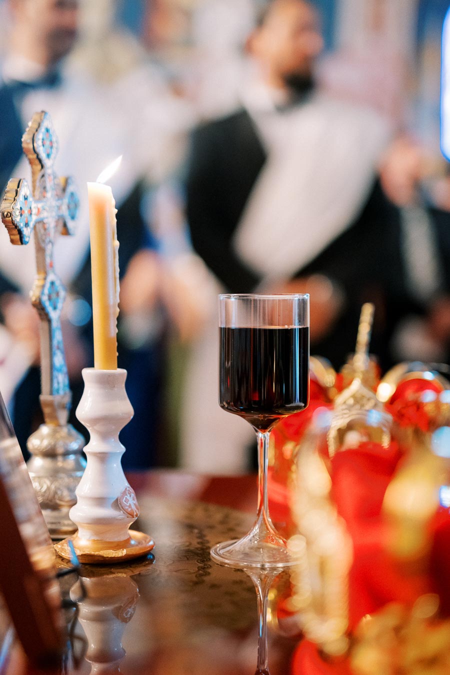 Close-up of a religious ceremony table featuring a lit candle, a decorative cross, and a glass of red wine, with blurred figures in ceremonial attire in the background