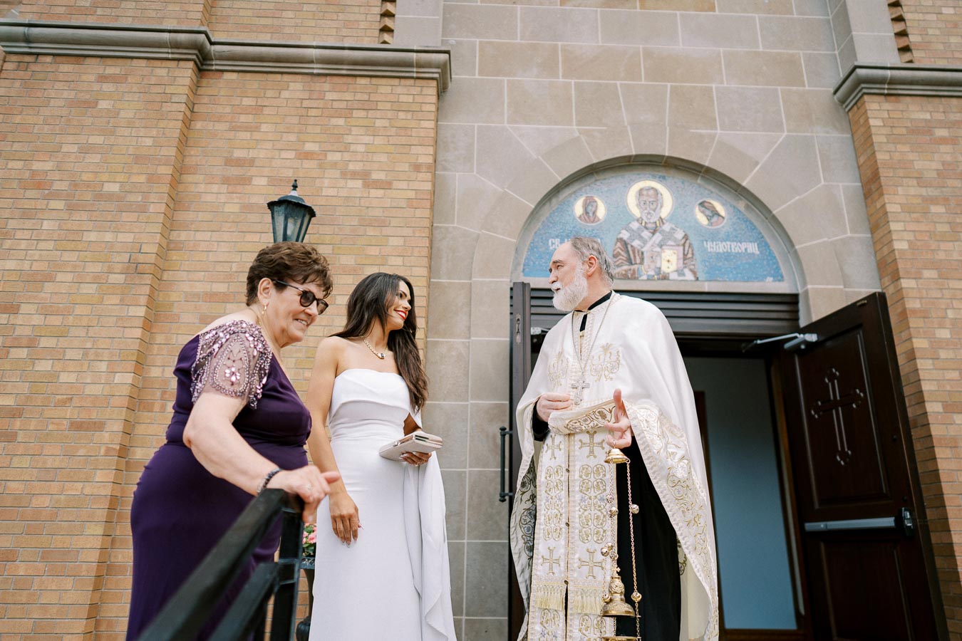Three people standing outside a church entrance, with one dressed in religious attire and two women in formal dresses, one holding a clutch, engaged in conversation.