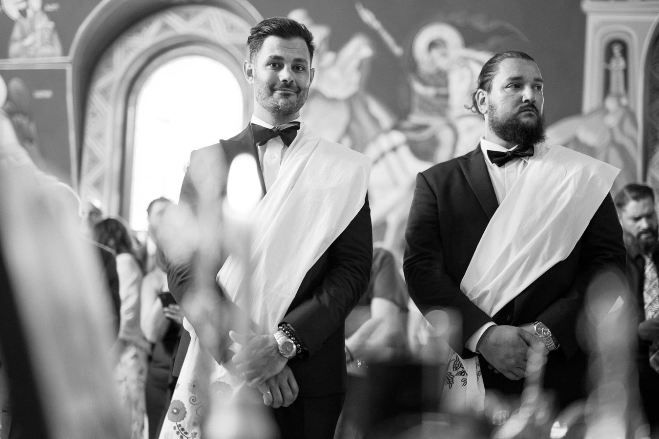 Black and white image of two men dressed formally in tuxedos and sashes, standing in an ornately decorated room with a large arched window in the background.