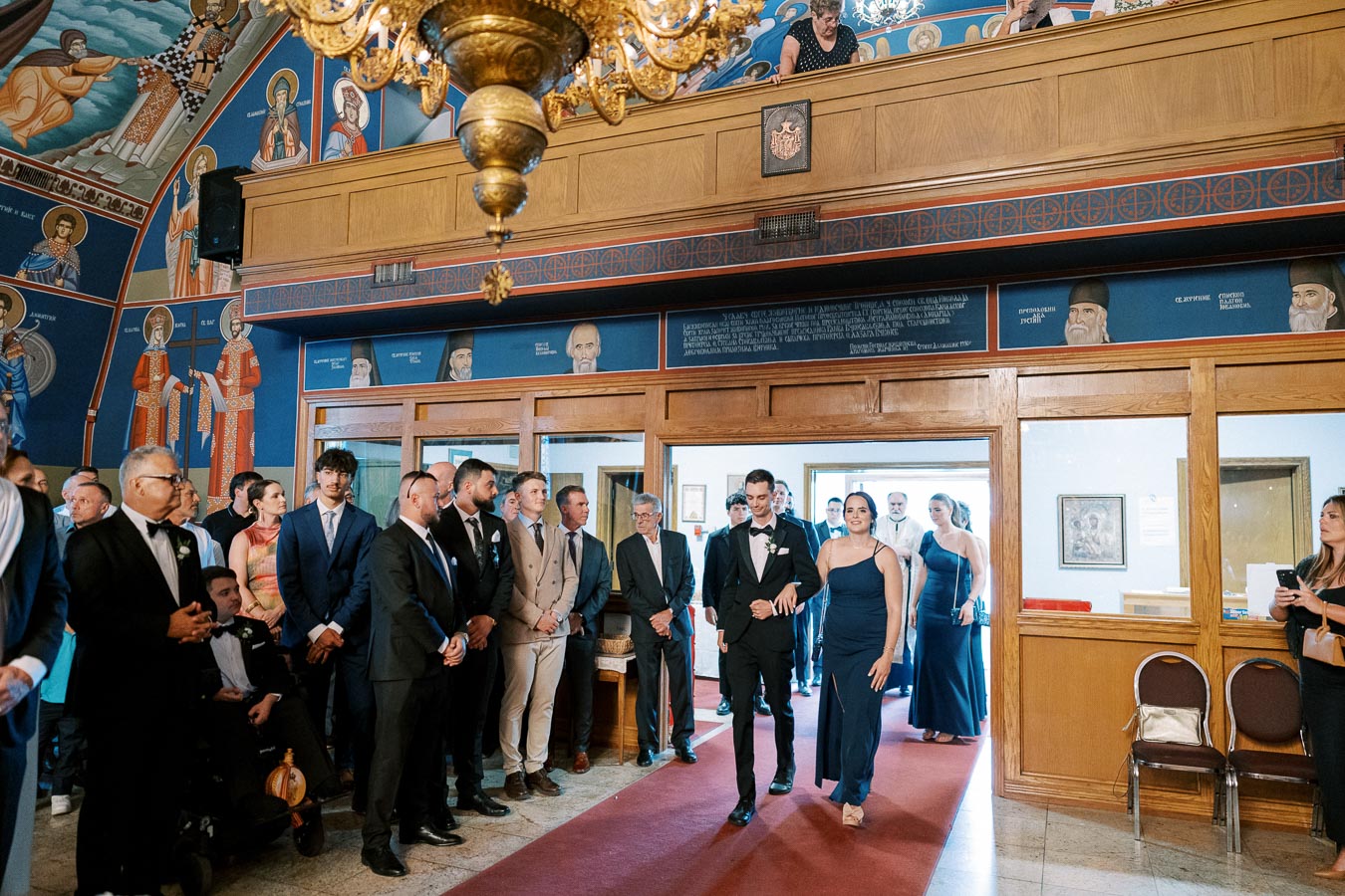 A wedding procession inside a beautifully decorated church, featuring guests dressed in formal attire and vibrant murals depicting religious figures on the walls. The atmosphere is elegant and celebratory, highlighting the cultural and spiritual significance of the event.