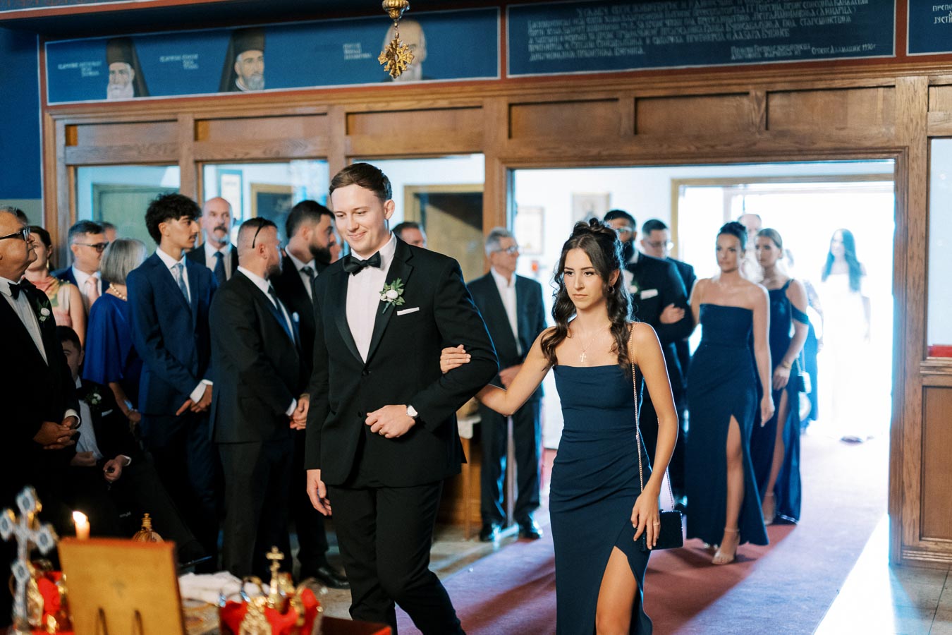 Elegant wedding procession with bridesmaid in navy dress leading groomsman in black tuxedo through a decorated church aisle, surrounded by guests
