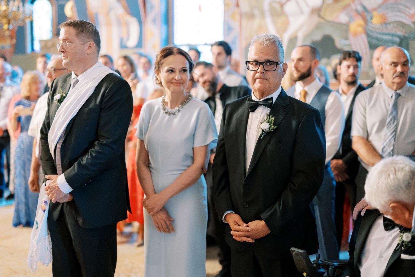 A group of well-dressed guests attending a formal event, with men in suits and a woman in a light blue dress, inside an elegantly decorated venue.