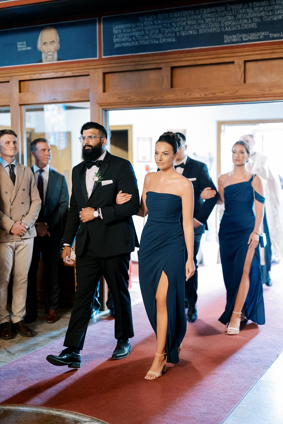 Elegant wedding procession with well-dressed bridal party, featuring men in suits and women in navy blue dresses, walking down a carpeted aisle in a formal setting.