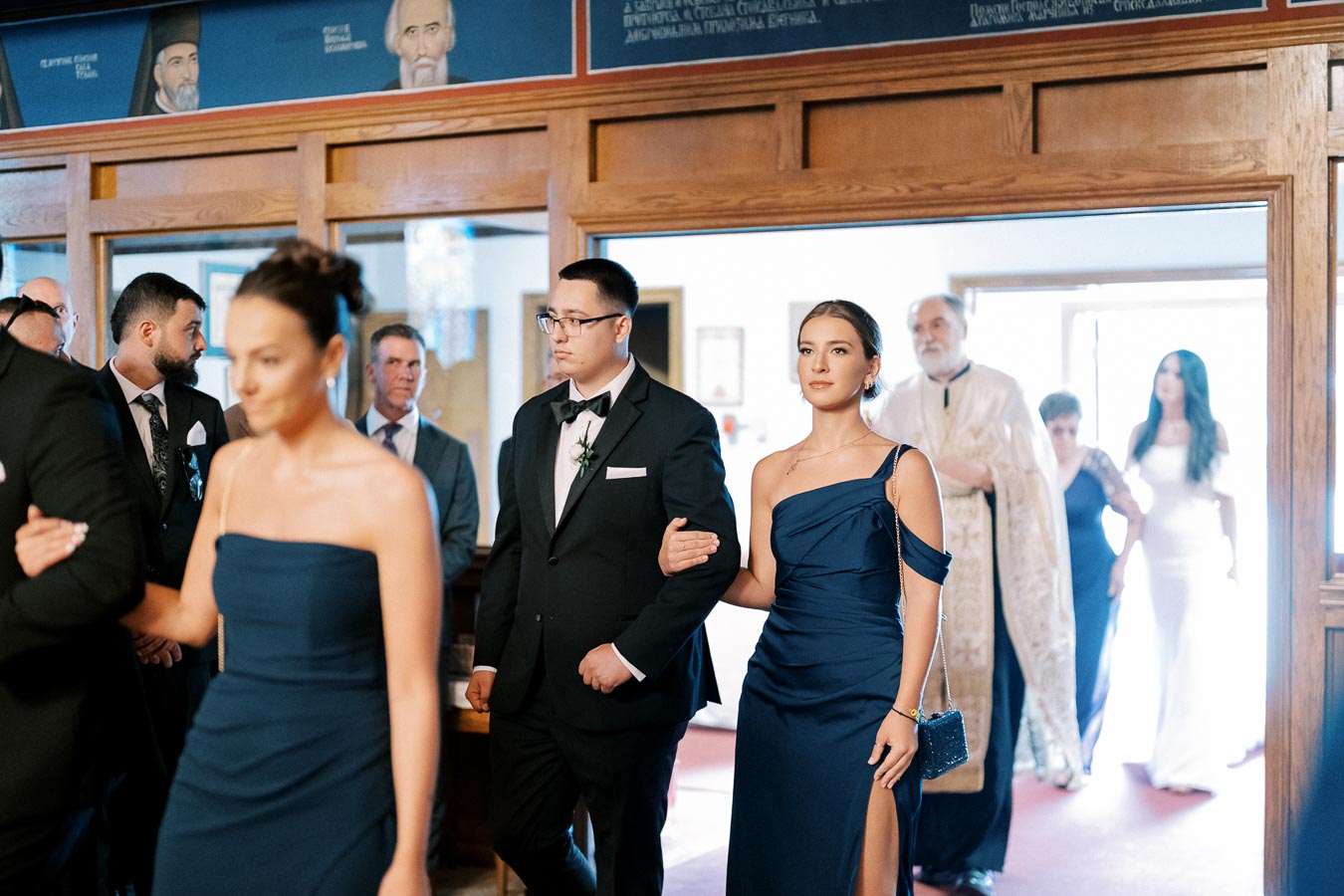 A group of formally dressed people walking in line during a wedding ceremony, with men in suits and women in elegant blue gowns, inside a decorated venue with wood-paneled walls.