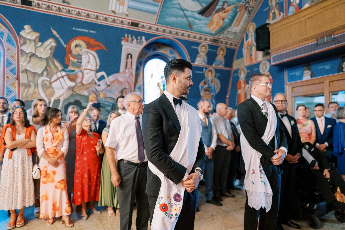Groom and best man wearing traditional sashes during a wedding ceremony in a church with vibrant religious murals, surrounded by guests in formal attire.