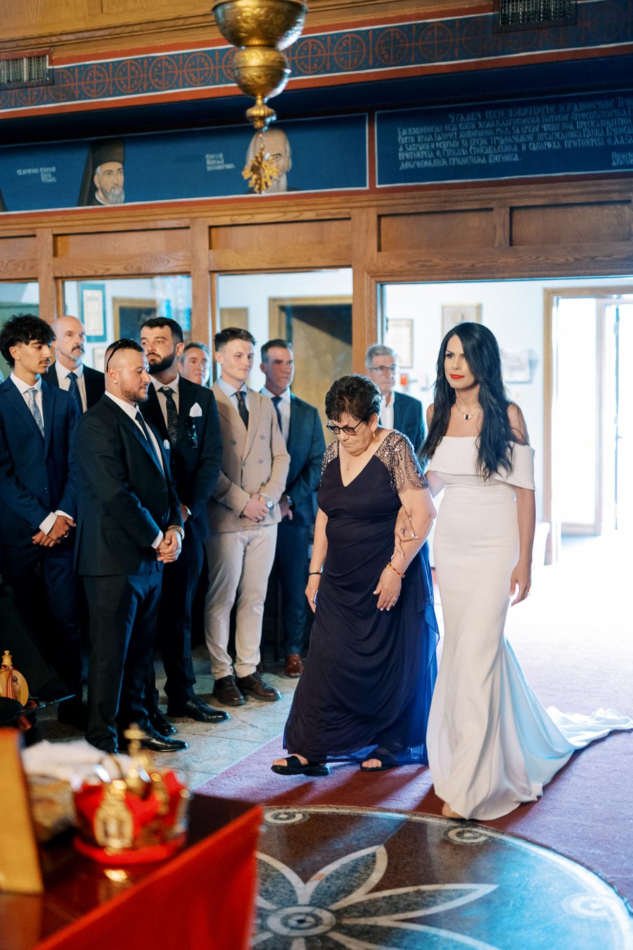 A woman in a white dress escorts an elderly woman in a navy gown down the aisle during a formal indoor ceremony, surrounded by elegantly dressed guests.