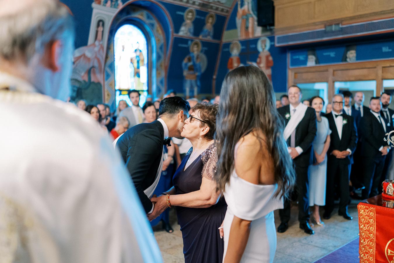 Wedding ceremony in a church with a groom in a black suit kissing an elderly woman in a purple dress, surrounded by guests and intricate stained glass windows in the background.