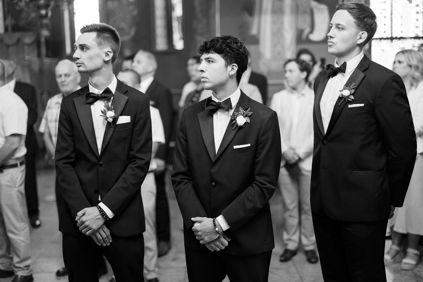 Three groomsmen wearing black tuxedos with bow ties and boutonnieres, standing indoors at a formal event, possibly a wedding ceremony.