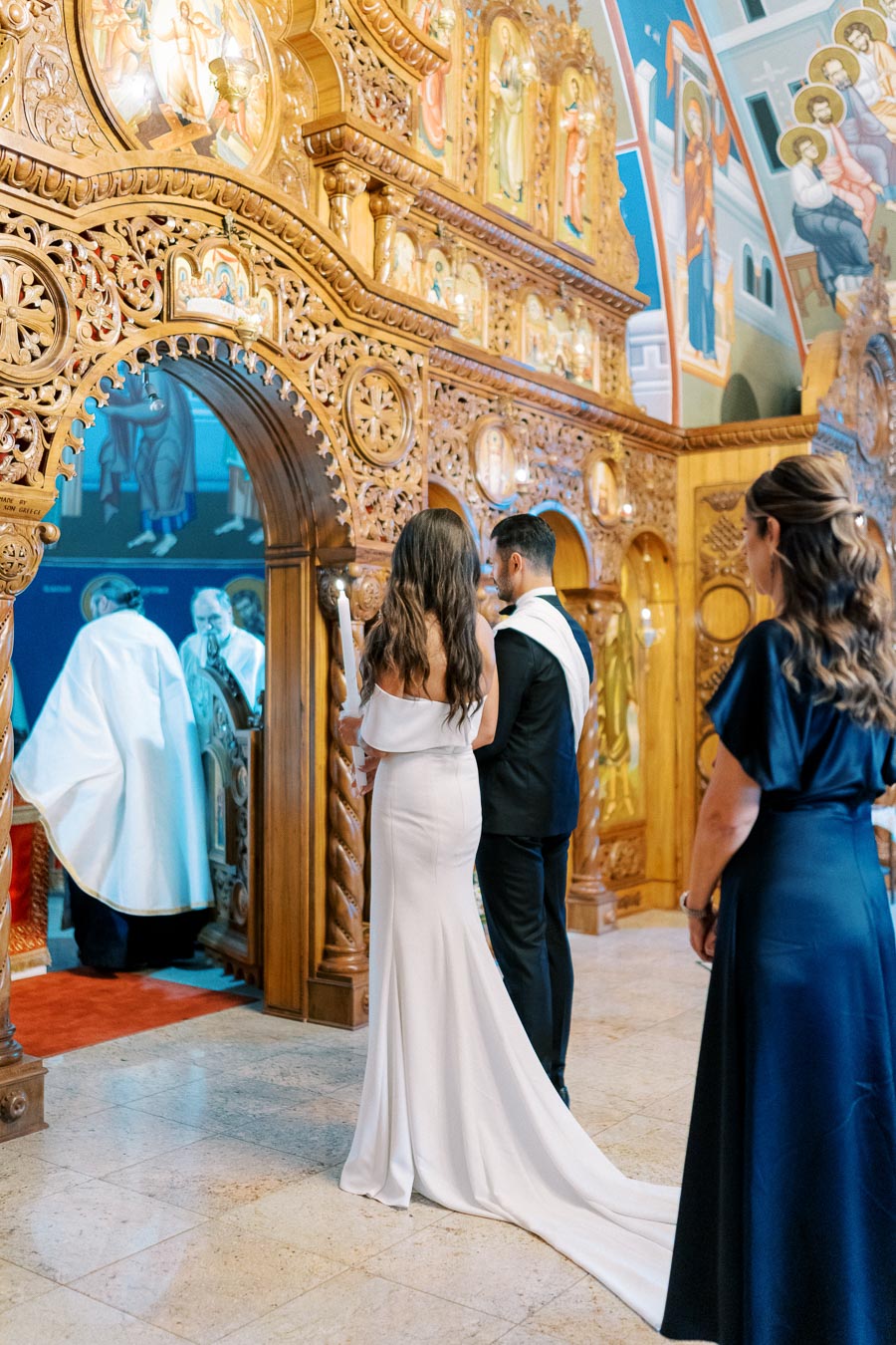 Bride and groom standing at the ornate entrance of a church during a wedding ceremony, with intricate religious carvings and icons in the background, capturing a solemn and beautifully decorated setting.