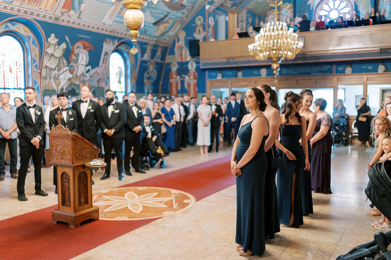 A traditional wedding ceremony inside a beautifully decorated church, with bridesmaids in elegant navy dresses standing in line and guests dressed in formal attire seated and standing in the background. Ornate chandeliers and vibrant religious murals adorn the walls and ceiling.