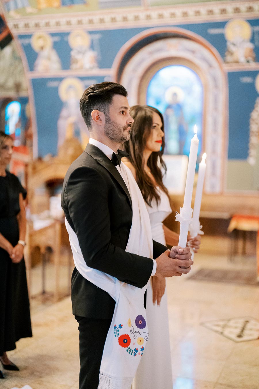 A couple standing together during a traditional wedding ceremony in a church, holding lit candles, with a backdrop of colorful religious icons and ornate architecture.